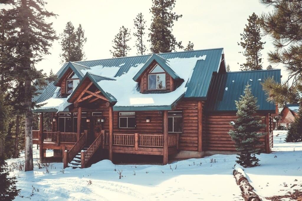 A snow covered log cabin in the forest surrounded by pine trees. 