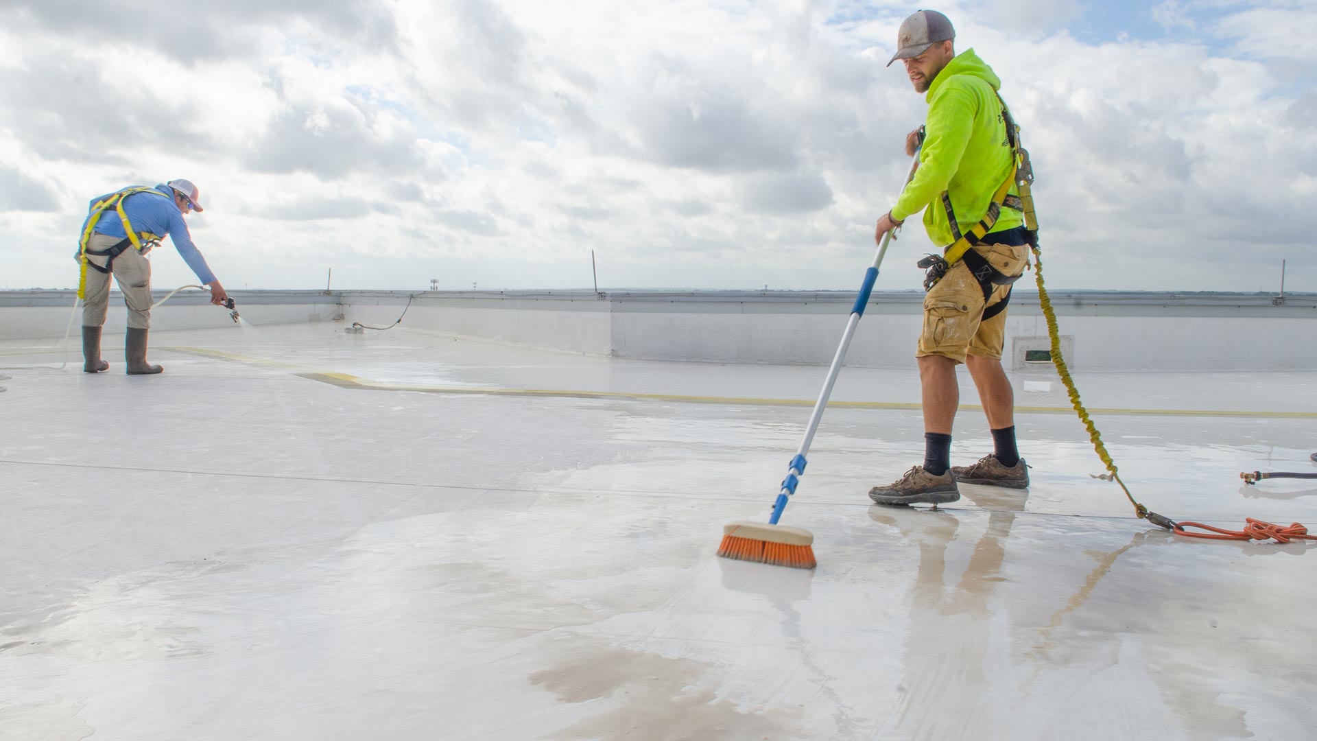 Roofing professionals cleaning a TPO flat roof with a broom and water.